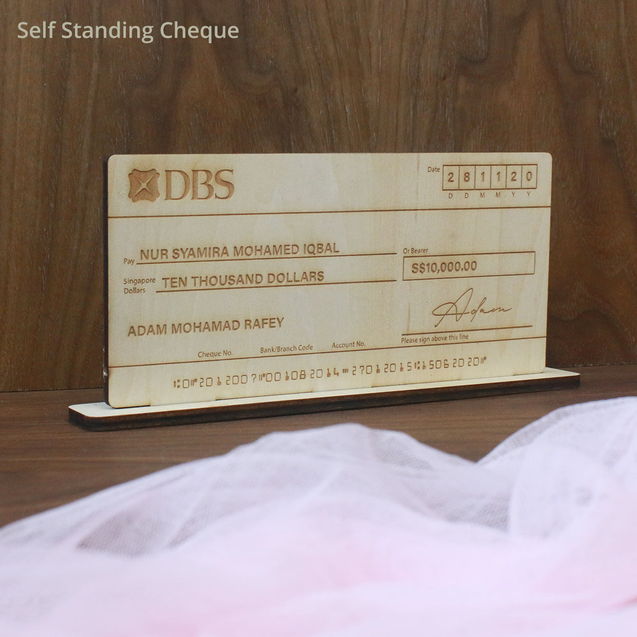 A light wood mock-up cheque with engraved text and signature, resembling a real bank cheque, displayed on a wooden surface.
