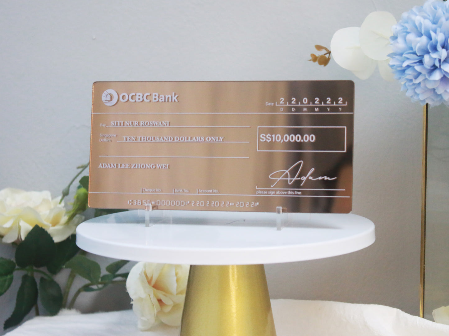 An acrylic mock cheque with bronze mirror finish, white printed text, displayed on a white stand with decorative elements around it.