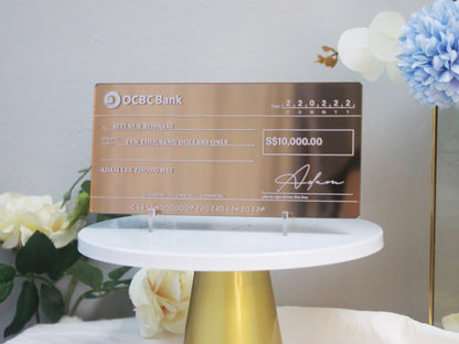 An acrylic mock cheque with bronze mirror finish, white printed text, displayed on a white stand with decorative elements around it.