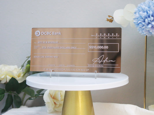 An acrylic mock cheque with bronze mirror finish, white printed text, displayed on a white stand with decorative elements around it.