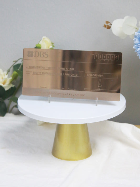 An engraved acrylic mock cheque with bronze mirror finish displayed on a white stand, with a gold base, against a floral backdrop.