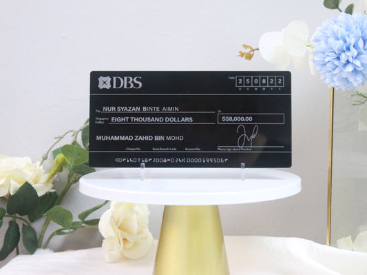 A glossy black mock cheque with white printed text, displayed on a white stand with decorative flowers in the background.