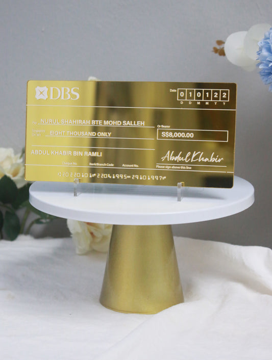 A gold mirror acrylic mock cheque with white printed text, displayed on a stand, against a backdrop with floral decorations.