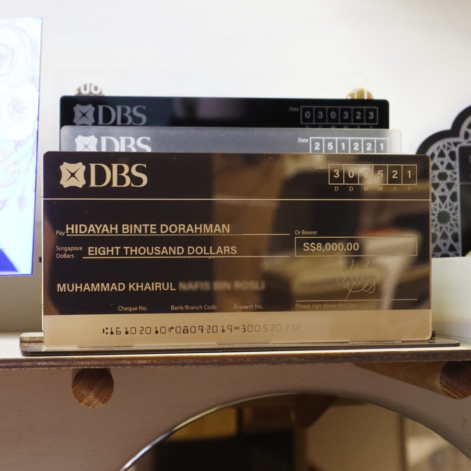A mock cheque displayed on a clear acrylic stand, with the bank name DBS visible on the top edge.