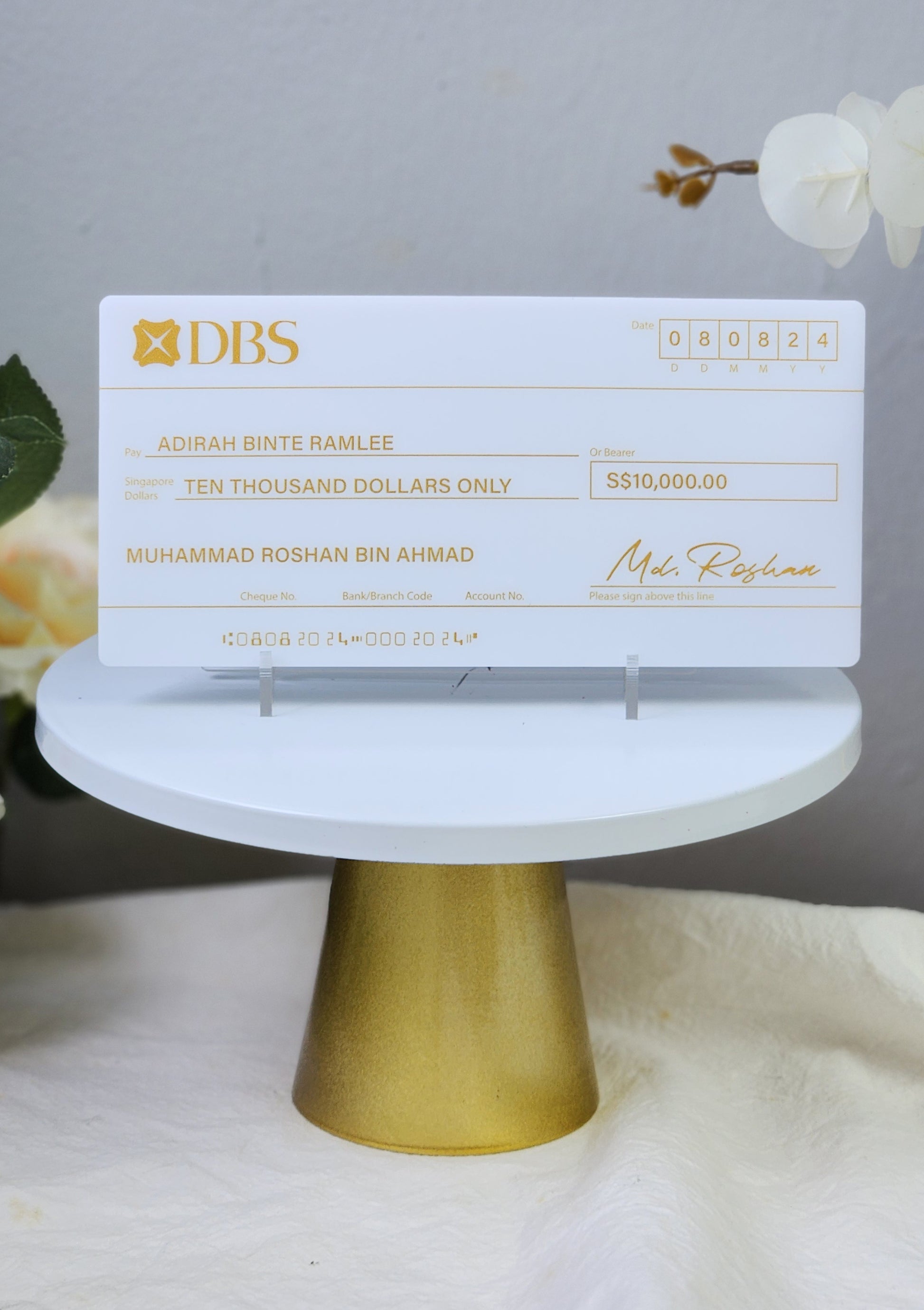 A customized mock cheque with gold printed text on opal white acrylic, displayed on a gold stand, with part of the text redacted for privacy.