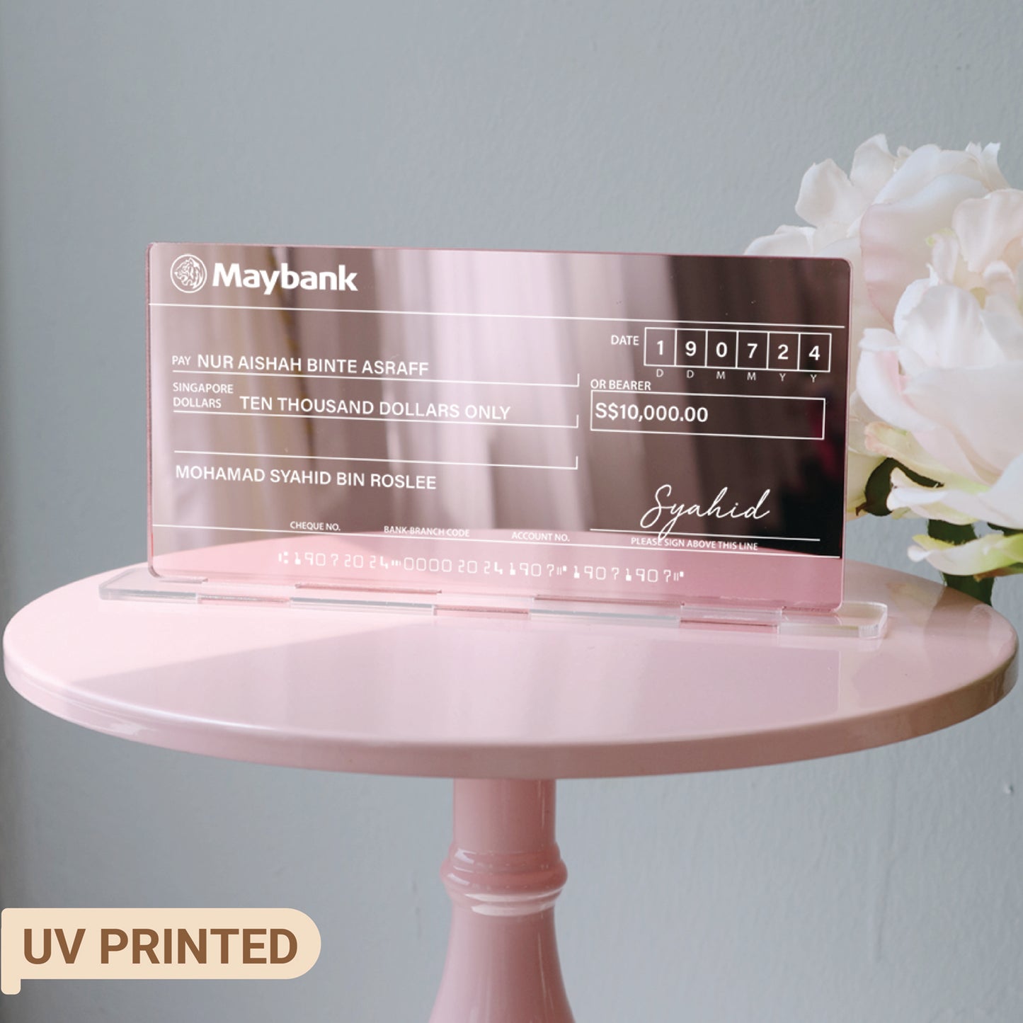 A pink mirrored acrylic mock cheque with white text, placed on a pink surface with a small wooden easel in the background.