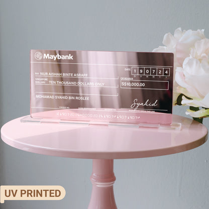 A pink mirrored acrylic mock cheque with white text, placed on a pink surface with a small wooden easel in the background.