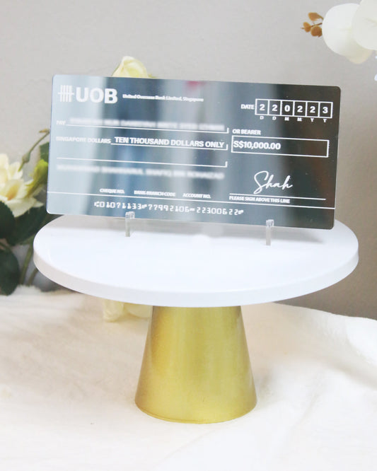 A mock cheque with white printed text on a silver mirror acrylic surface, presented on a white stand, resembling a banknote design with fields for personalization.