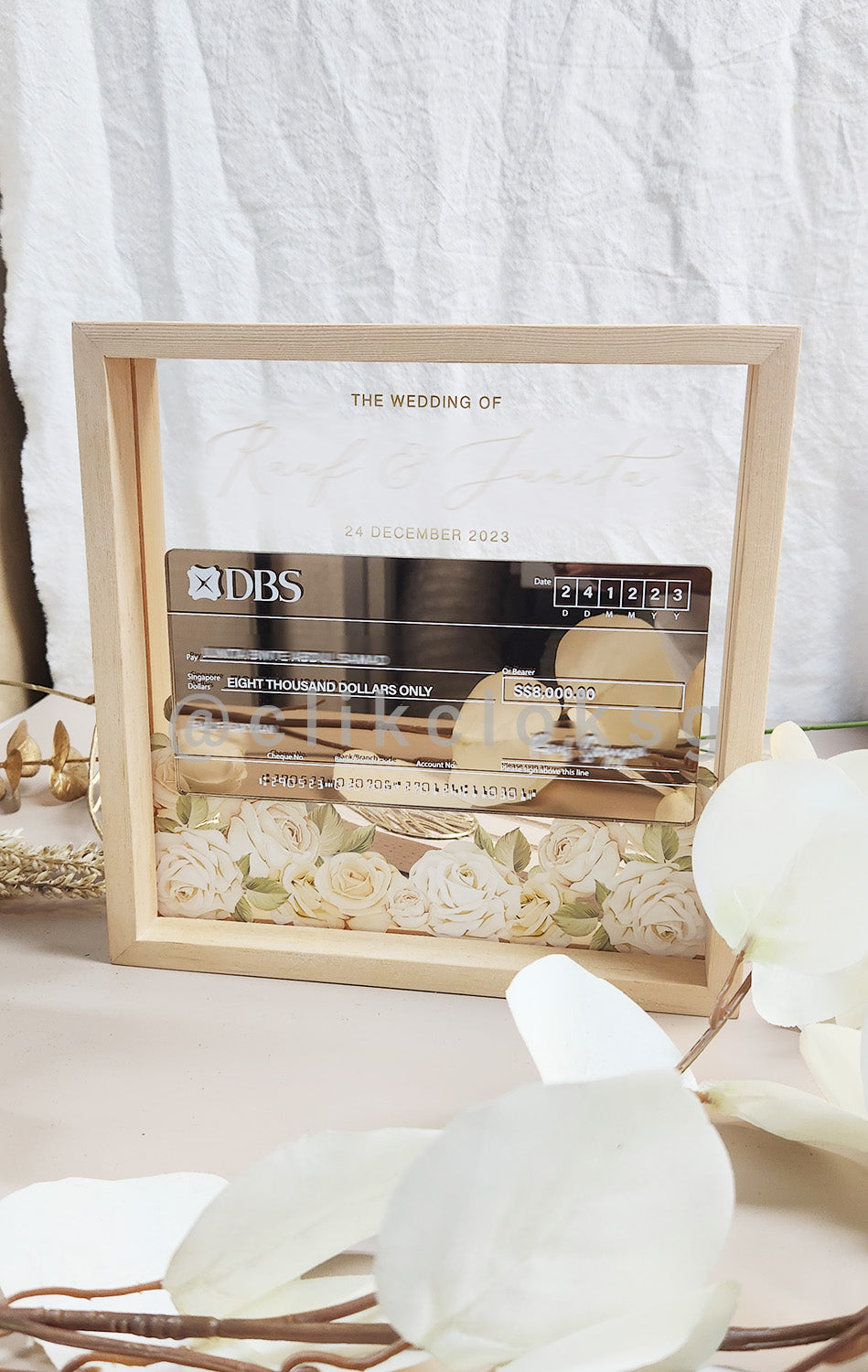 A mock cheque display with floral patterns in a pinewood frame, with a clear acrylic body and a custom pop-up cheque with customization options.
