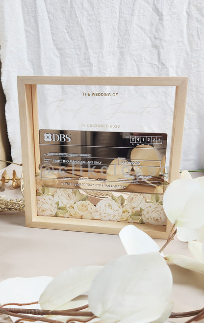 A mock cheque display with floral patterns in a pinewood frame, with a clear acrylic body and a custom pop-up cheque with customization options.