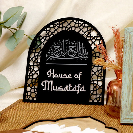 A black acrylic emblem with a lattice carve design and Arabic script that reads 'House of Mustafa'. It is positioned against a backdrop with a blue frame and decorative elements.