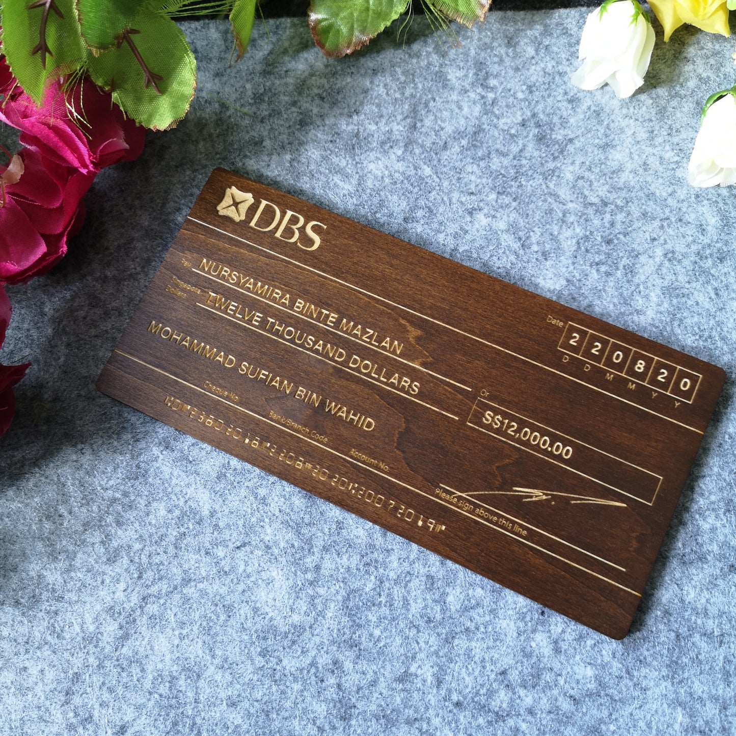 A customized mock-up cheque for a hantaran with dark wood finish displayed on a white stand, surrounded by floral decorations.